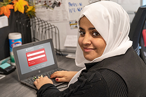 A student looks over her shoulder and smiles at the camera. She is typing on a laptop at her desk. 