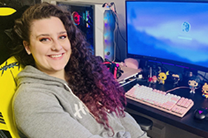 A systems engineer smiles in front of her home workstation with a computer screen and a pink keyboard.