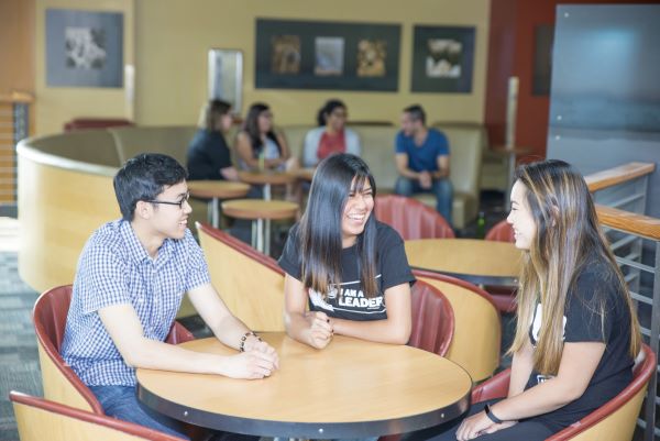 1 male and 2 female students sit at a table talking and smiling