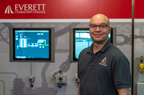 A male college instructor stands in front of industrial equipment and screens.