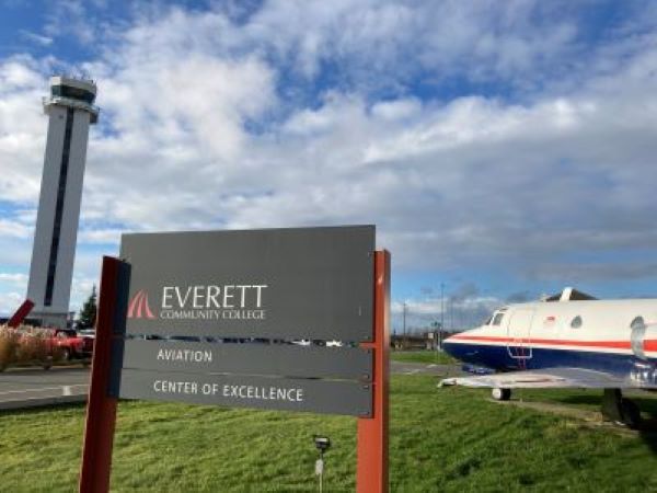 Everett Community College Featherstar Logo with Aviation and Center of Excellence signage in the foreground, air control tower in the left background, small plane aircraft on in the right background against a blue sky with white clouds, all situated on a green lawn next to asphalt under the tower