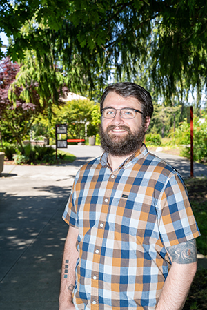   Individual standing on a sunlit pathway in a park, smiling at the camera. They are wearing a checked shirt and have tattoos on their arms.