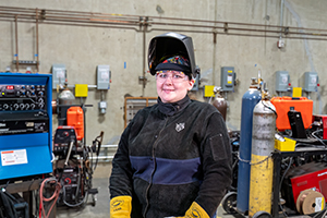 AMTEC Welding Student Erika Manweiler, wearing welding gear and a face mask protector lifted up to reveal her face, posing in a workshop environment.