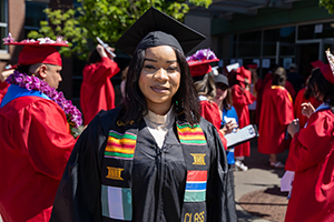 Graduate, Fatou Jaiteh, in cap and gown, adorned with a colorful stole, smiling at a college commencement ceremony, surrounded by other graduates in red gowns.