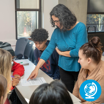An instructor works with a group of students in a classroom, with an icon of a globe in the corner.