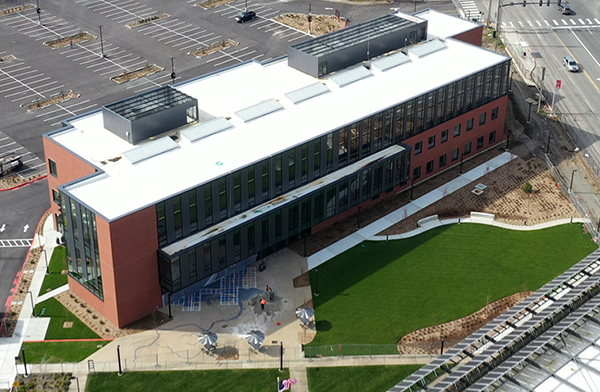 an aerial photo shows a large brick and glass three story building with a parking lot behind, a wide street to the right and a grassy courtyard in front.