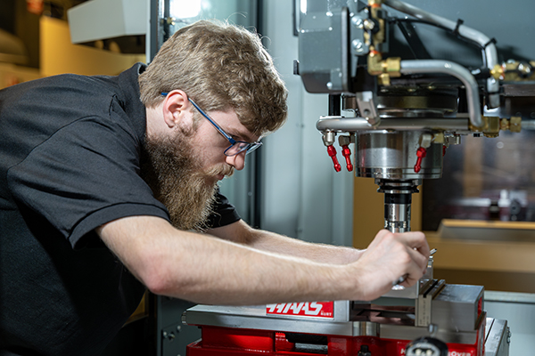 A focused man with a beard and glasses operates a metal milling machine, wearing a black shirt. The setting is industrial, with a serious tone.