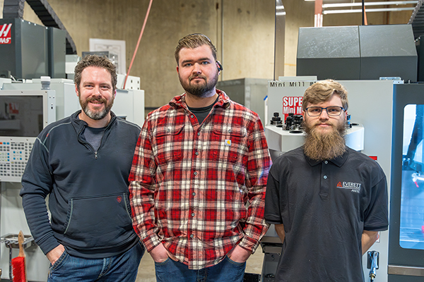 Three men stand side by side in a workshop, smiling at the camera. They are dressed casually. The setting features industrial machinery, conveying teamwork.