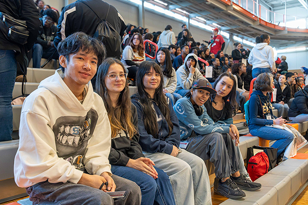 Five students look toward the camera while they sit on bleachers in a packed gymnasium.