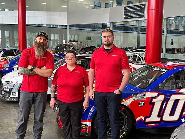 Three students in red shirts stand in front of a row of race cars.