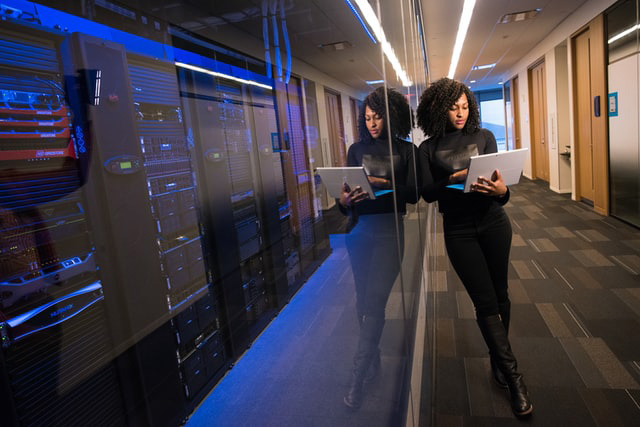 Woman with a computer stands near a bank of computer servers.