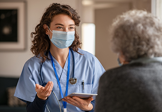 Female healthcare professional wearing a facemask speaks with an older woman. 