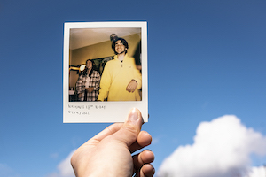 Elise McIntyre- photograph of a poloroid of a man in a yellow hoodie held up in front of a blue sky.