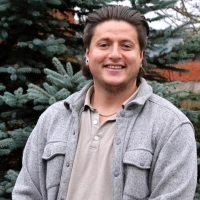 A photo of Zac Lounsbury wearing a tan polo, a grey sweater, and medium length brown hair.